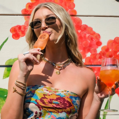 A vibrant image of a woman enjoying a popsicle, dressed in a floral top and accessorized with layered gold necklaces and bracelets. Her sunglasses and smile highlight her playful summer vibe.