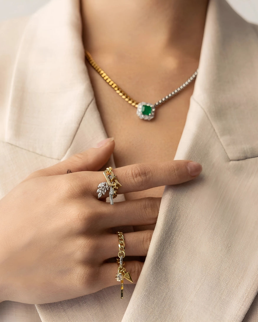 Close-up of a model wearing "The Emerald Pendant Necklace," a gold and silver chain necklace with a green emerald centerpiece, paired with layered gold charm rings, including "The Lightning Bolt Charm Ring" and "The Star Charm Ring.