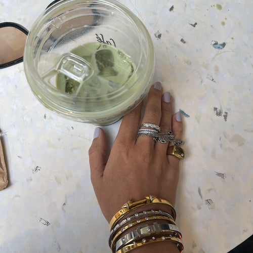 A hand adorned with multiple gold and silver stacked rings, paired with chunky bracelets, gently holding a glass of iced matcha latte. The jewelry pieces include a dainty diamond ring, a statement band, and a bangle, styled against a soft, minimalistic background of textured white fabric.