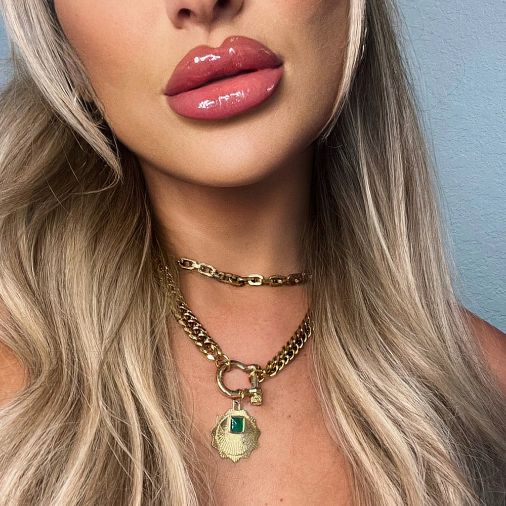 A close-up shot of a woman wearing bold chain necklaces in gold and silver, paired with a striking emerald pendant. Her glossy lips enhance the luxurious look.

