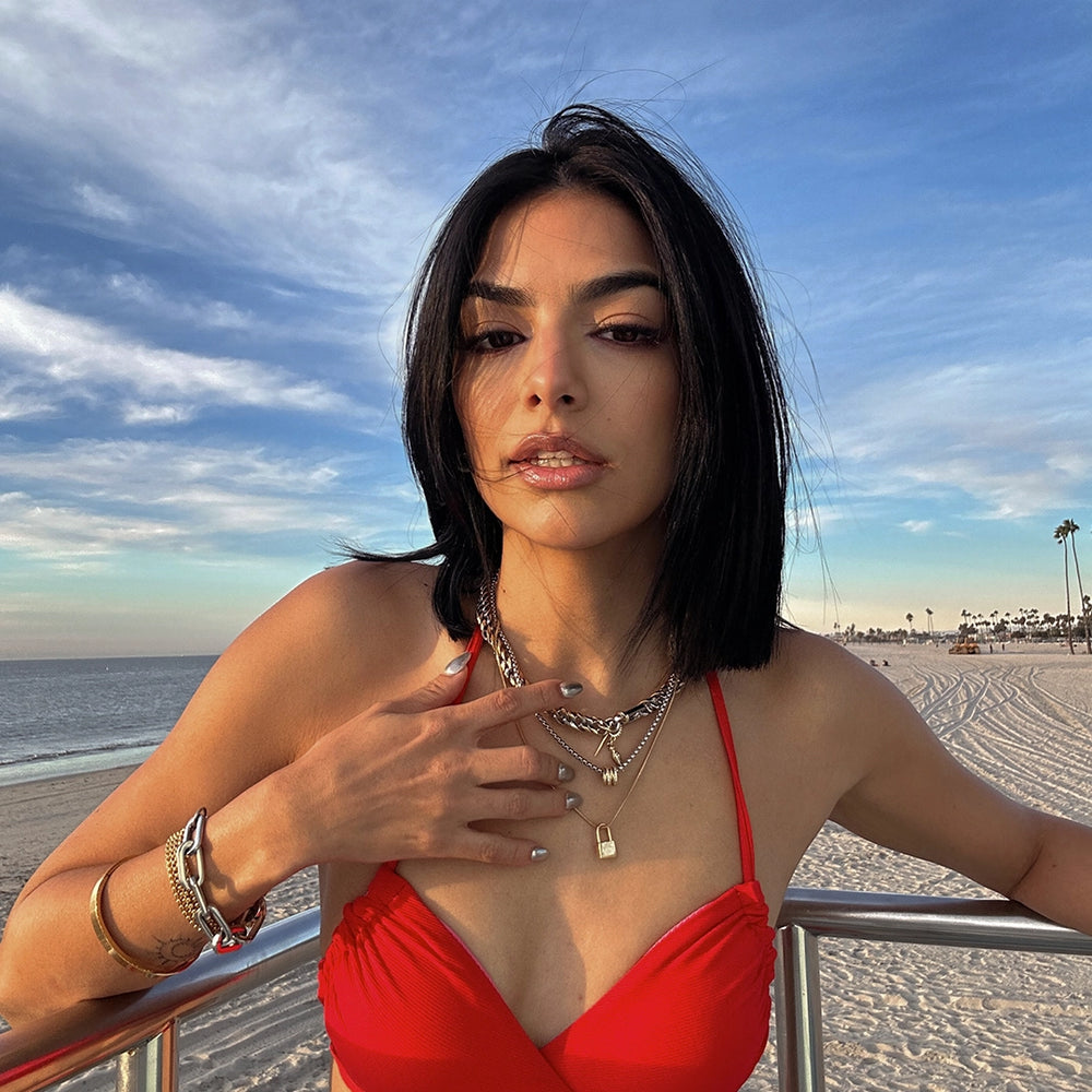 A woman in a red swimsuit, layered with Artizan Joyeria’s bold gold necklaces and bracelets, posing against a serene beach sunset