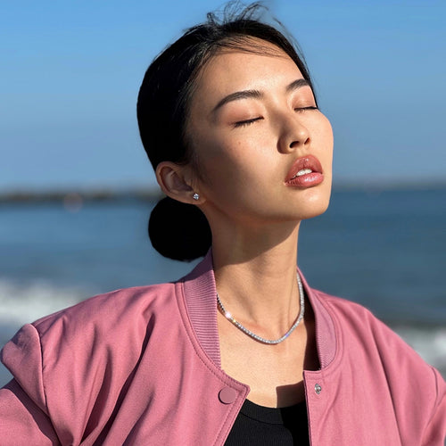 A serene woman in a pink jacket and black top, wearing a sparkling Artizan Joyeria tennis necklace, basking in the sunlight by the ocean