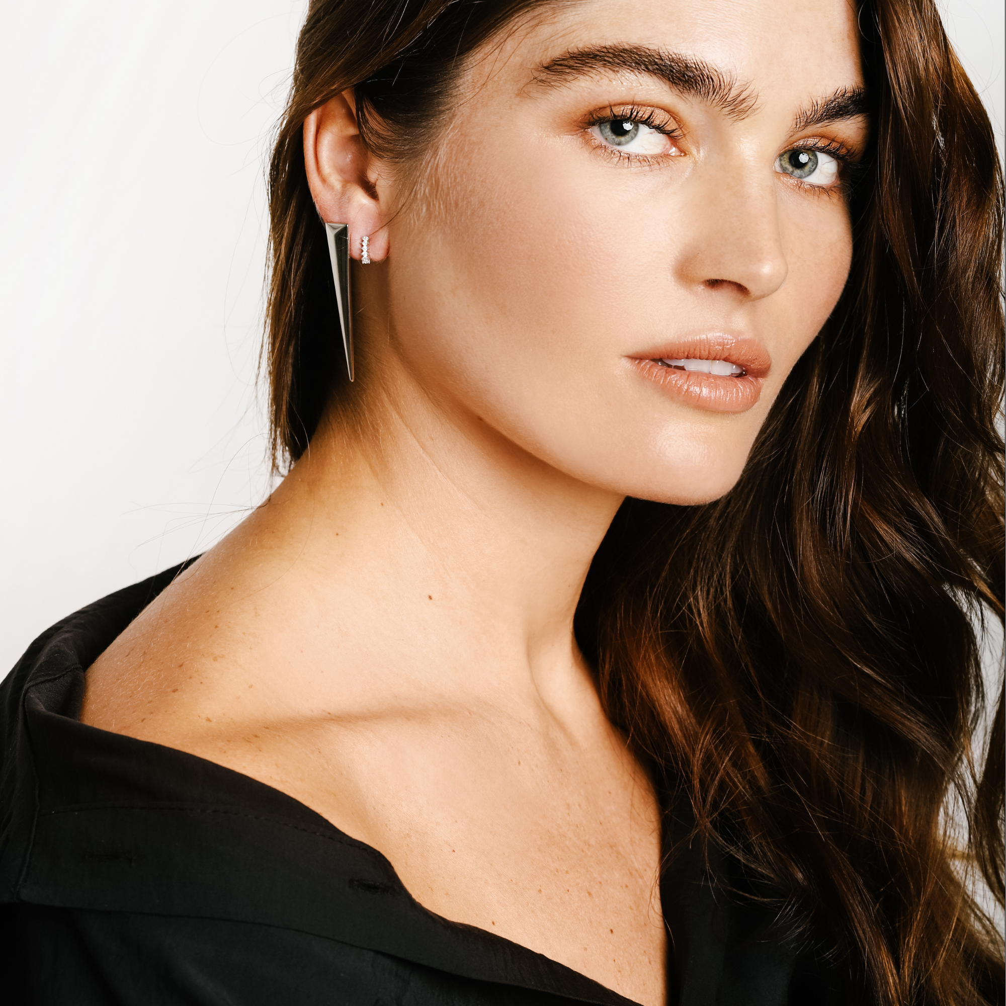 Model wearing the Needle earring, 925 sterling silver which is a long triangle earring. She is also wearing a silver huggies earring (size: 0.9 mm) with cubic zirconia. ||| SILVER