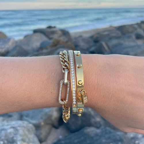 Close-up of a wrist adorned with multiple gold bracelets, including chain, studded, and charm designs, with an ocean background. The bracelets convey a luxurious and summery vibe