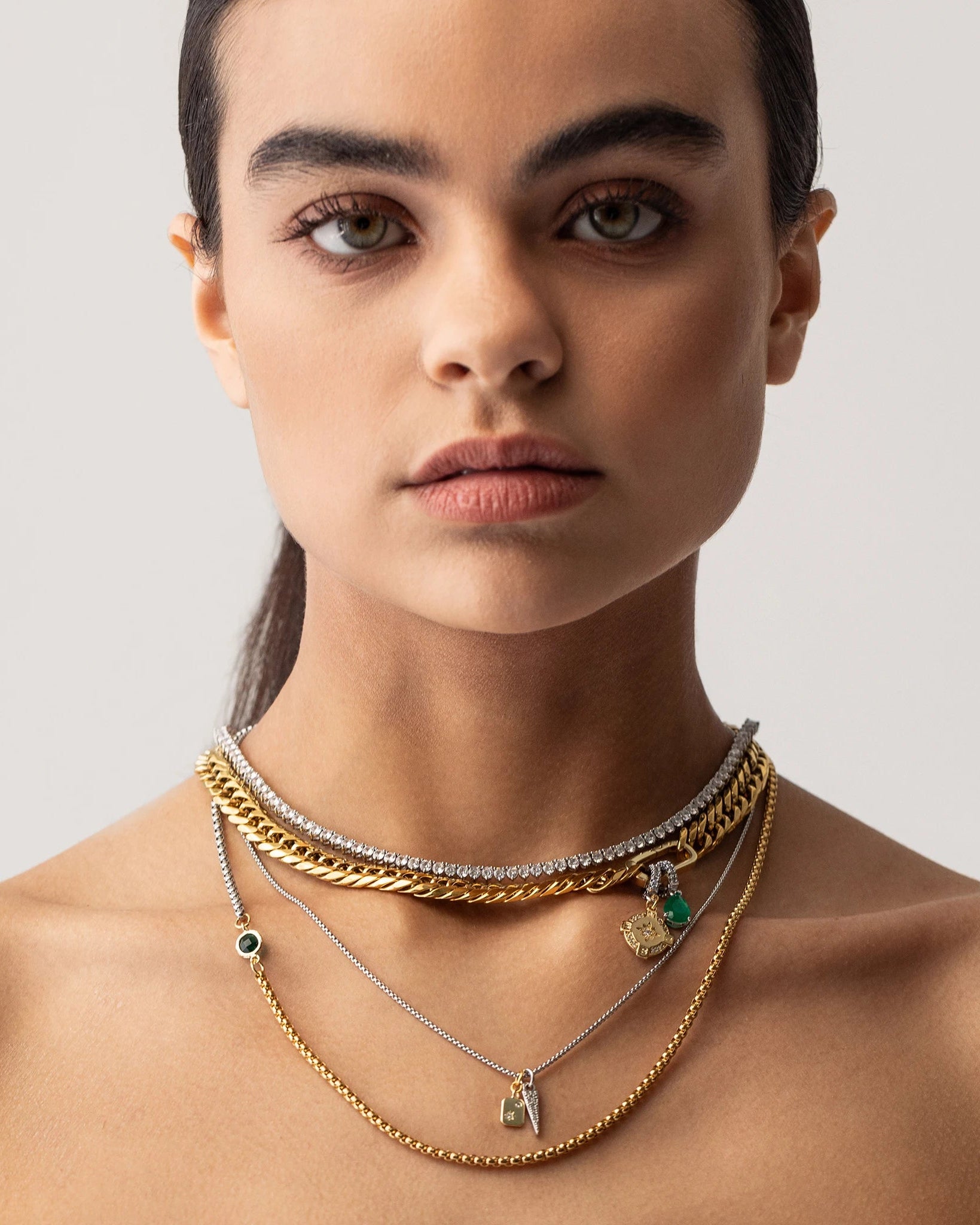 Model wearing The Just Click Emerald Tennis Necklace Set, a four-piece gold and silver necklace set featuring a variety of chains with diamond-like accents, emerald accents, and a variety of charms.