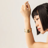 Model wearing the Queen's Crown Bracelet Stack, a four-piece gold bracelet stack featuring bangles with a variety of accents, including the word "ARTIZAN," emerald accents, and a textured design.