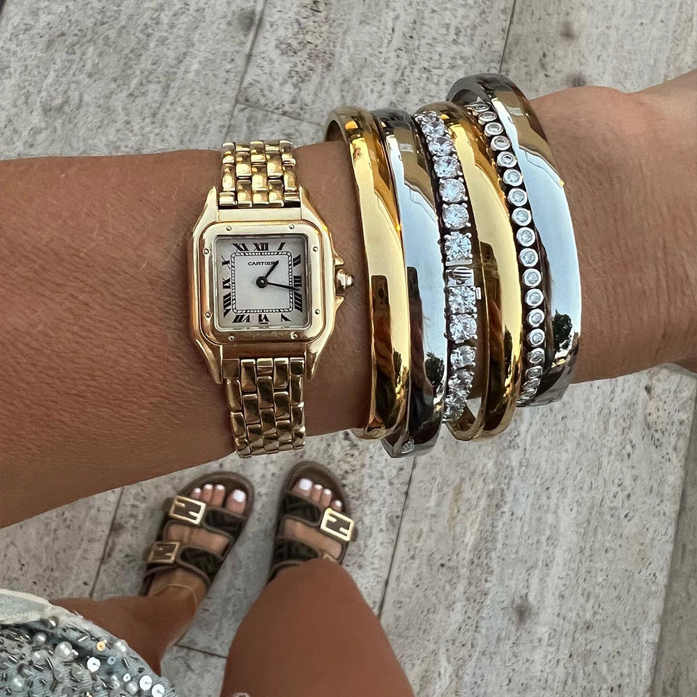 This image shows a close-up of a person's wrist adorned with several luxurious bracelets of Artizan Joyeria and a gold wristwatch. The gold watch has a square face with Roman numerals and a metallic band. Alongside the watch, the person is wearing three sleek bangles: one gold, one silver, and another encrusted with diamonds. The individual is dressed in casual yet stylish attire, visible from the bottom half, wearing designer sandals with gold buckles and standing on a stone-tiled surface. 