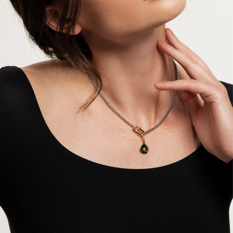 The Box Charm Necklace, which is a single-piece mixed metal necklace featuring a silver chain with a gold carabiner clasp and a black and gold teardrop charm with a star detail.