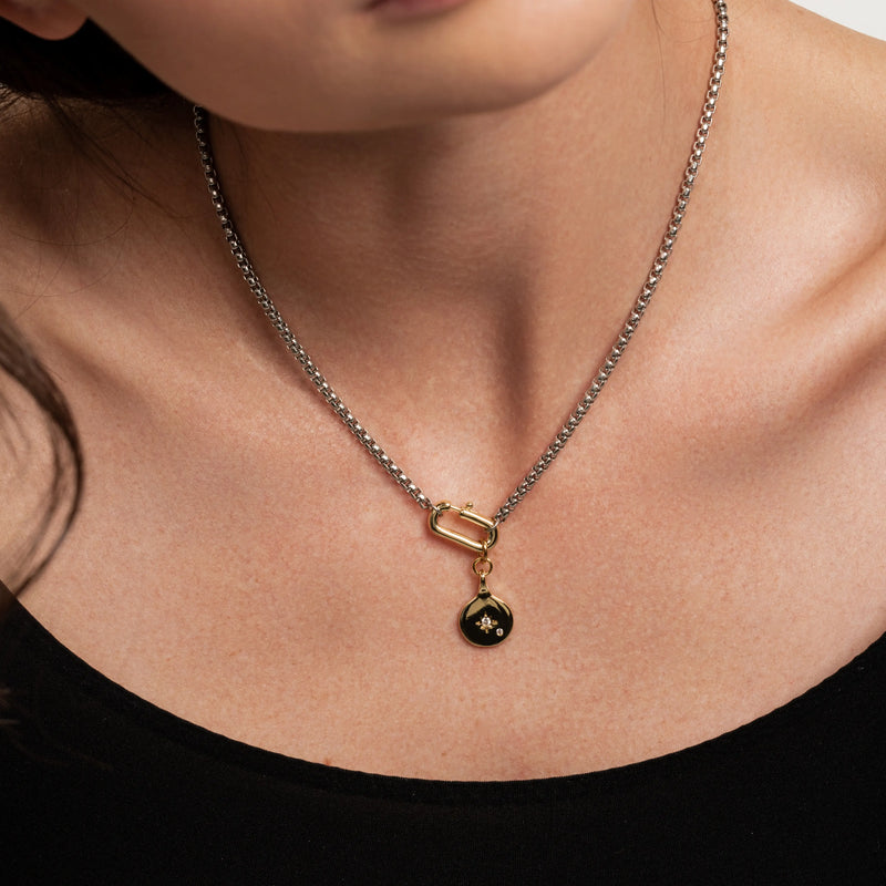 The Box Charm Necklace, which is a single-piece mixed metal necklace featuring a silver chain with a gold carabiner clasp and a black and gold teardrop charm with a star detail.