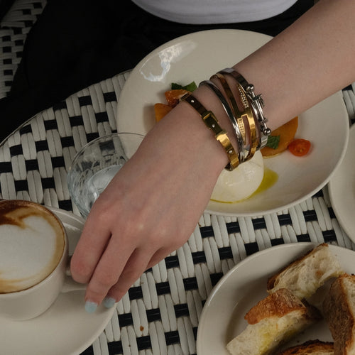 A woman's wrist wearing a stack of gold and silver bracelets, a gold bangle bracelet with a geometric pattern, and a silver cuff bracelet with a bold design all of them from artizan  joyeria . The woman is sitting at a table with a cappuccino, a plate of burrata cheese, and a basket of bread.