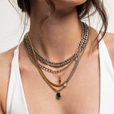 The Camaleon Layered Necklace Set, which is a layered necklace set featuring 5 pieces: a gold chain with a star pendant, a silver chain with a green stone pendant, a silver and gold chain mix, and a gold chain with a spike pendant, worn by a woman in a white top.