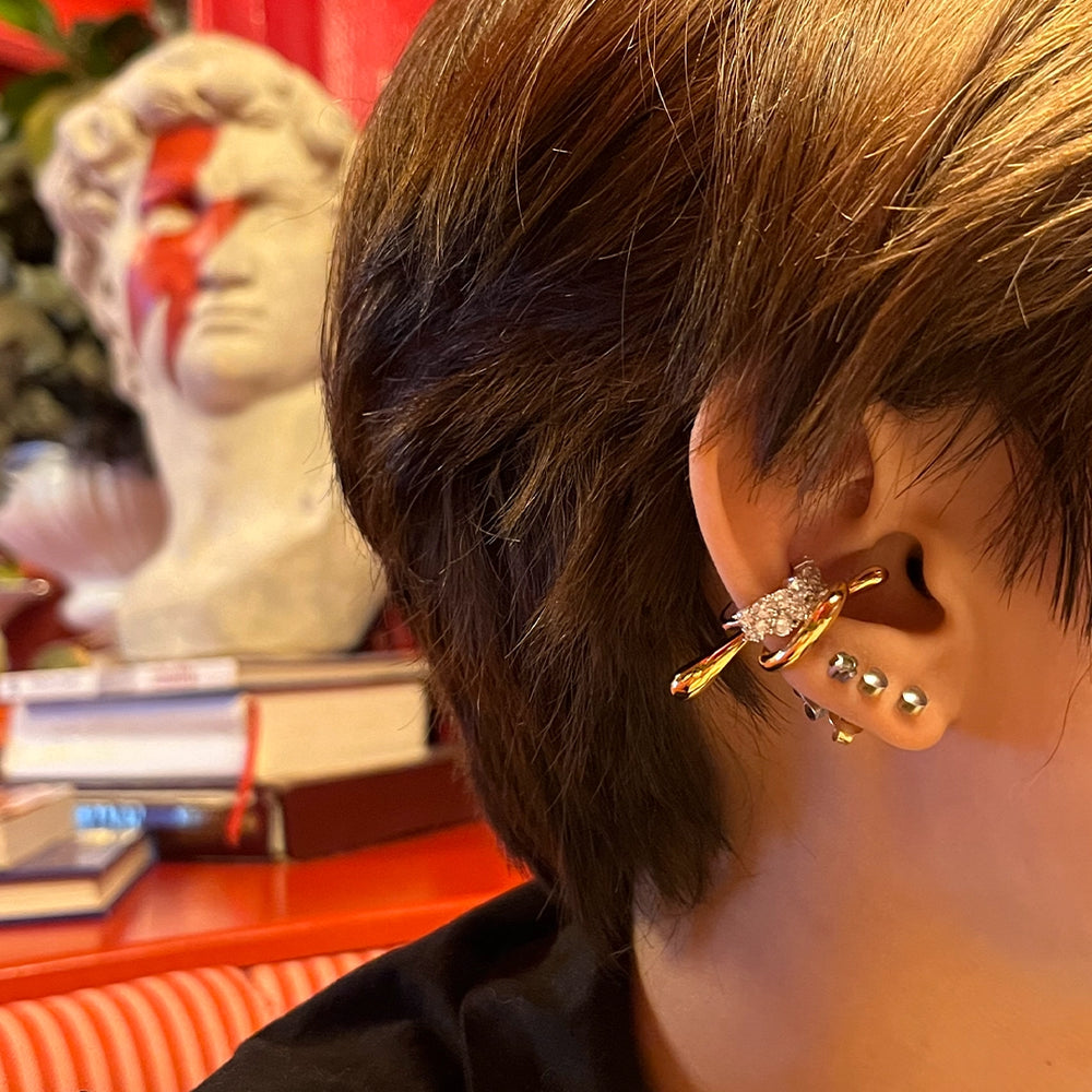 A close-up of a person's ear adorned with multiple piercings, including a diamond-studded MAGNIFICENT ear cuff and several small silver studs. The person has short brown hair, and in the background, there is a white bust sculpture painted with a red lightning bolt, resembling David Bowie’s iconic Aladdin Sane look