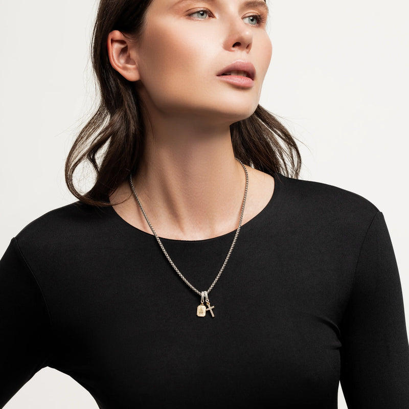 The Double the Charm Necklace, which is a single silver-toned chain necklace featuring two distinct charm pendants—a lock and a cross—styled for a bold, modern look. 