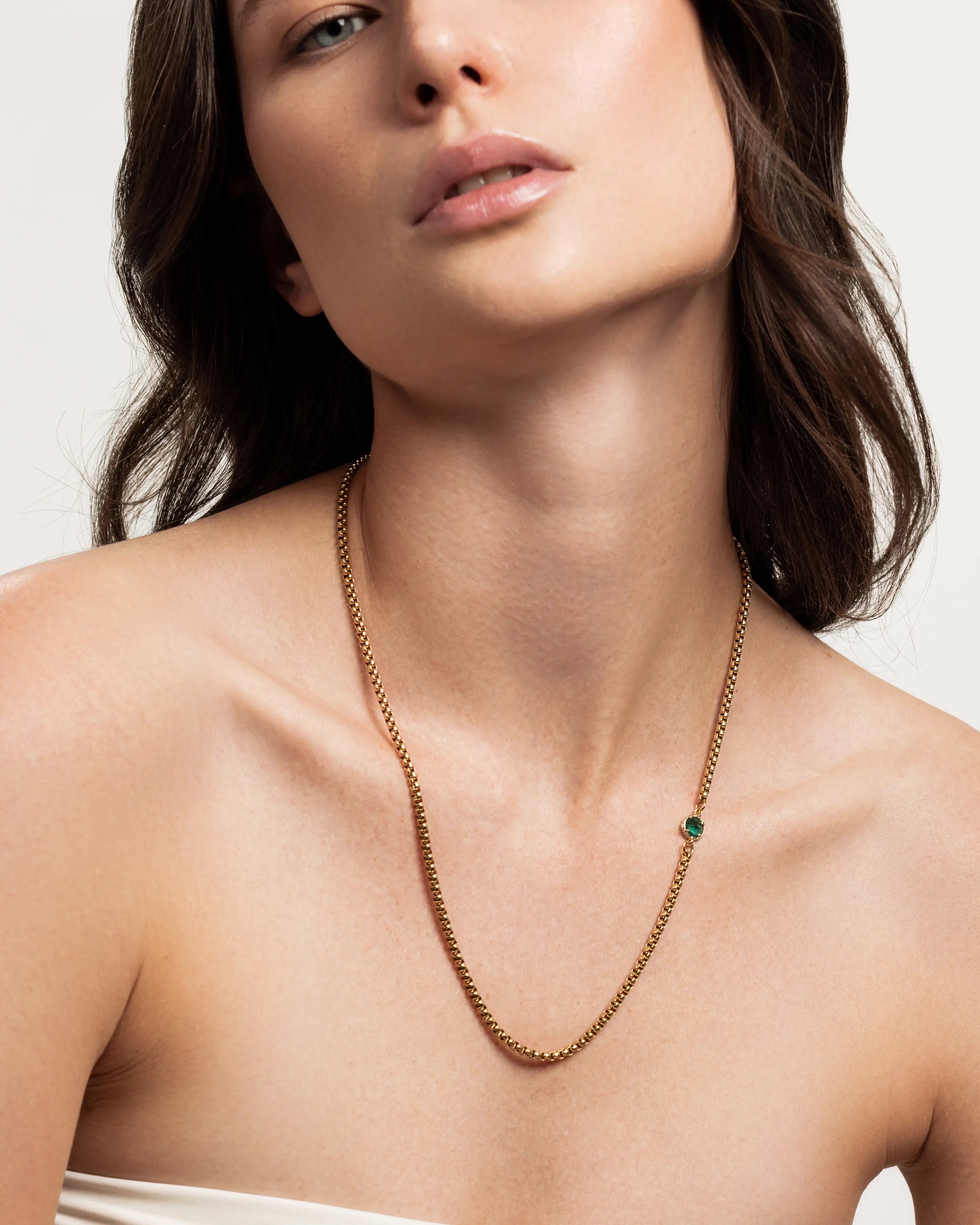 The Emerald Point Necklace, which is a delicate gold chain necklace featuring a single emerald stone, worn by a woman in a strapless top. ||| GOLD