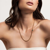 The Emerald Point Necklace, which is a delicate gold chain necklace featuring a single emerald stone, worn by a woman in a strapless top. ||| GOLD