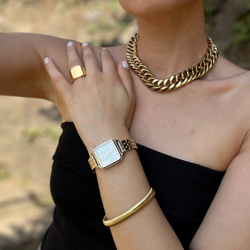 This image features a person wearing bold gold Artizan's jewelry. The individual is adorned with a chunky GOLD CLEO NECKLACE, a square-shaped FOREVER RING GOLD, a sleek gold CATERPILAR BANGLE , and a square gold wristwatch with a metallic band.Is dressed in a black, strapless top, and the overall style is elegant, with the gold jewelry making a strong, luxurious statement. The pose highlights the accessories, with the person resting one hand on their shoulder and the other near their chest.