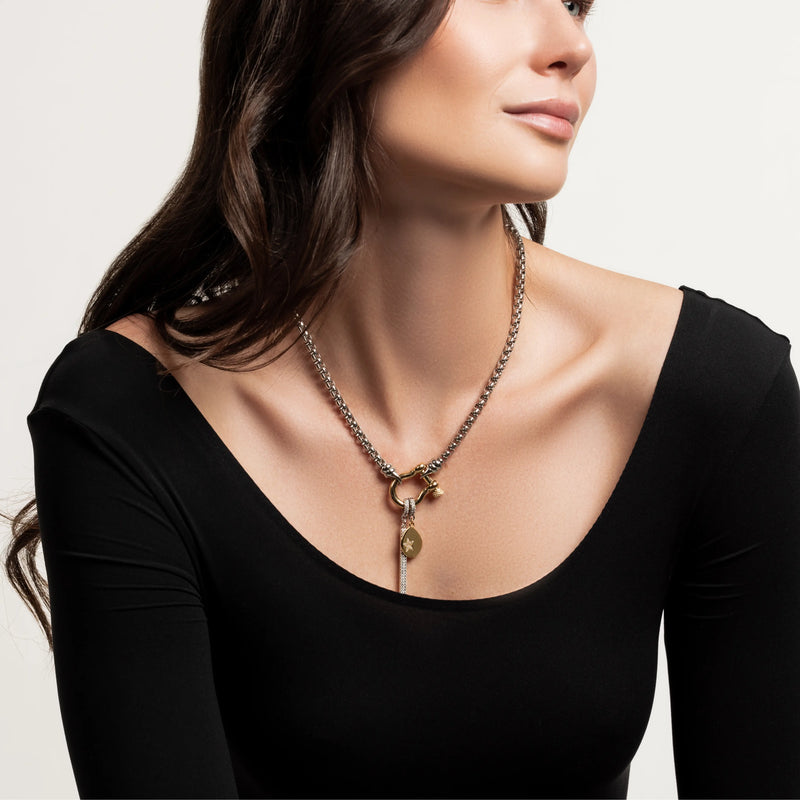 The Herradura Signature Double Take Gold, which is a mixed metal necklace featuring a gold and silver chain with a toggle clasp, adorned with a gold charm and a key pendant, worn by a woman in a black long-sleeve top. 
