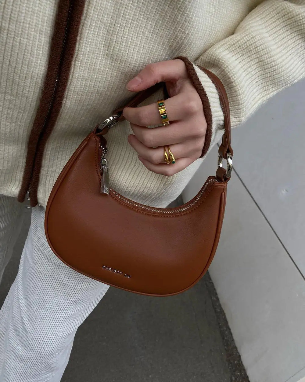 A close-up of a hand holding a brown handbag, wearing a mix of silver and gold rings, including a chunky ring with colorful accents. The hand is dressed in a cozy beige sweater with a zipper.