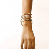 Model wearing the Khanti Bracelet Stack, a five-piece gold and silver bracelet stack featuring bangles with a variety of  accents, including a knotted bangle, a buckle bangle with diamond-like stones, a bar bangle, and a bangle with diamond-like stones and a heart charm.