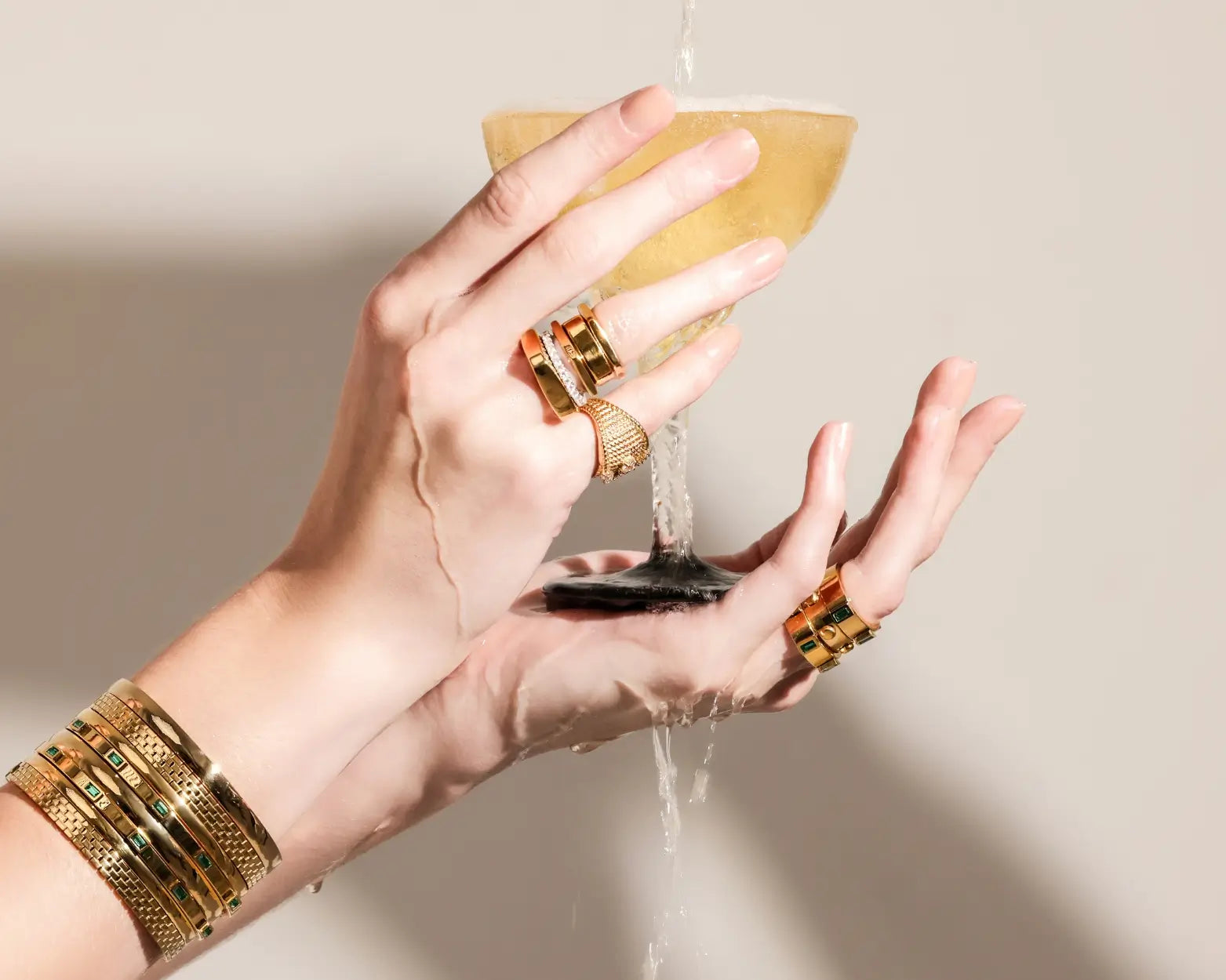 Hands holding a champagne glass under running water, styled with chunky gold rings and stacked gold bangles with emerald and pavé accents.