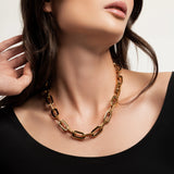 The Puerto Chain Necklace Gold, which is a bold gold chain necklace featuring large, rectangular links, worn by a woman in a black top.
