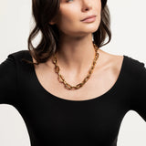 The Puerto Chain Necklace Gold, which is a bold gold chain necklace featuring large, rectangular links, worn by a woman in a black top. ||| GOLD