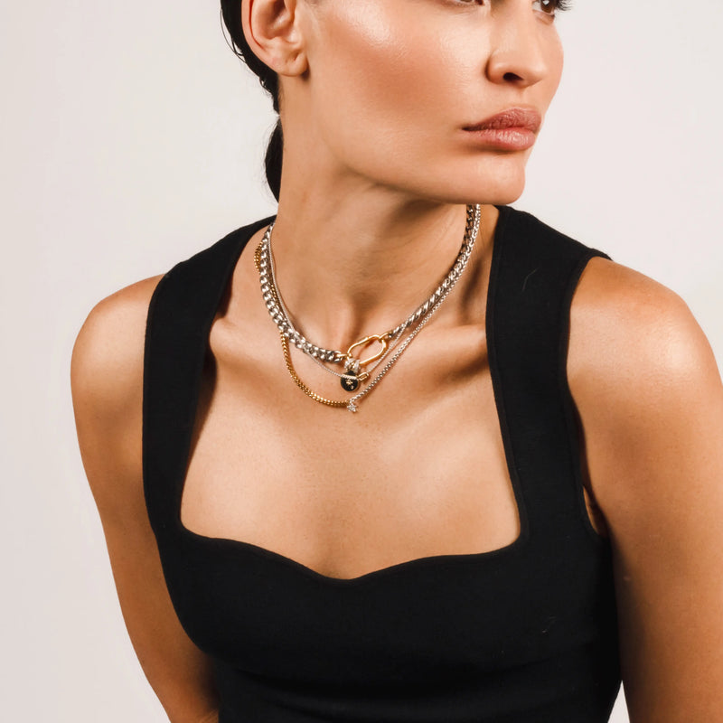 The Sleek Carabiner Cosmic Layered Necklace Set, which is a 3-piece mixed metal necklace set featuring chunky silver and gold chains, a gold carabiner clasp, and celestial-inspired charms for a modern, layered look.