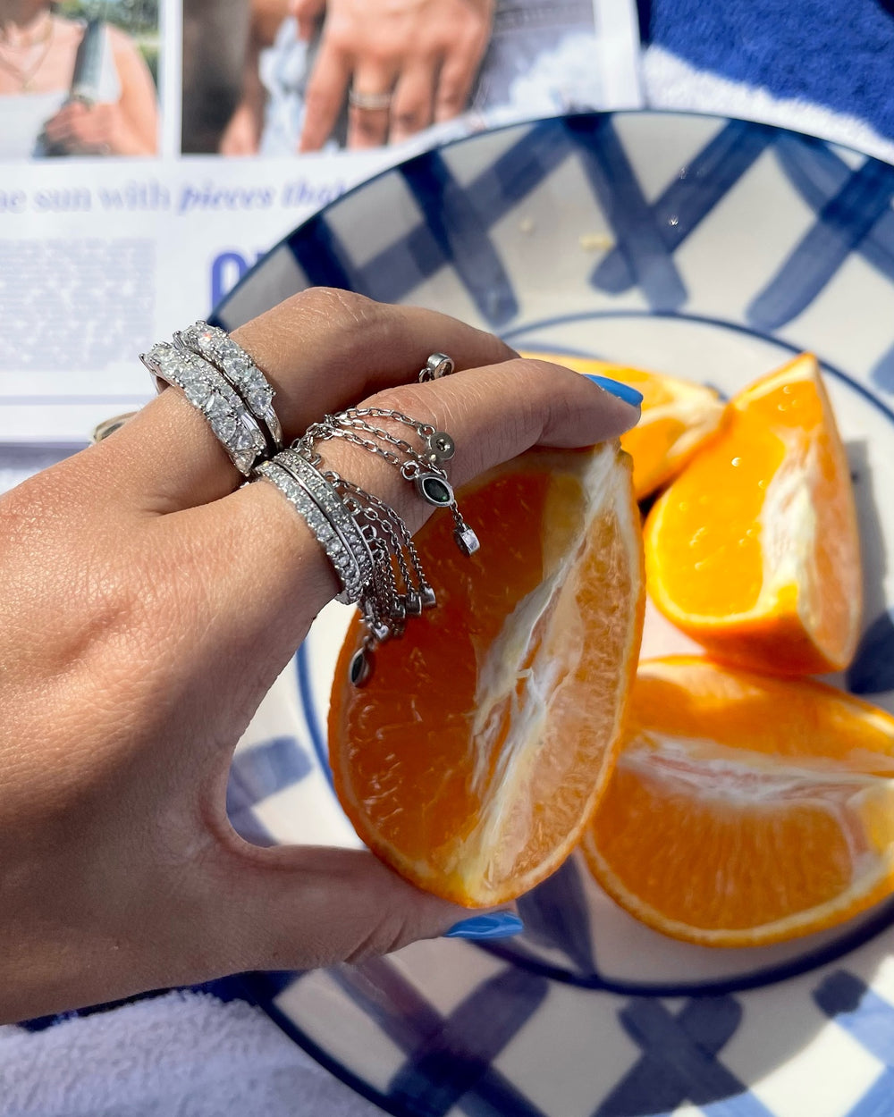 A woman's hand with several silver Artizan joyeria rings on each finger, holding a slice of orange. The rings are a mix of styles, including a chunky cocktail ring, a delicate chain ring, and a stack of thin bands. The orange is on a blue and white checkered plate.