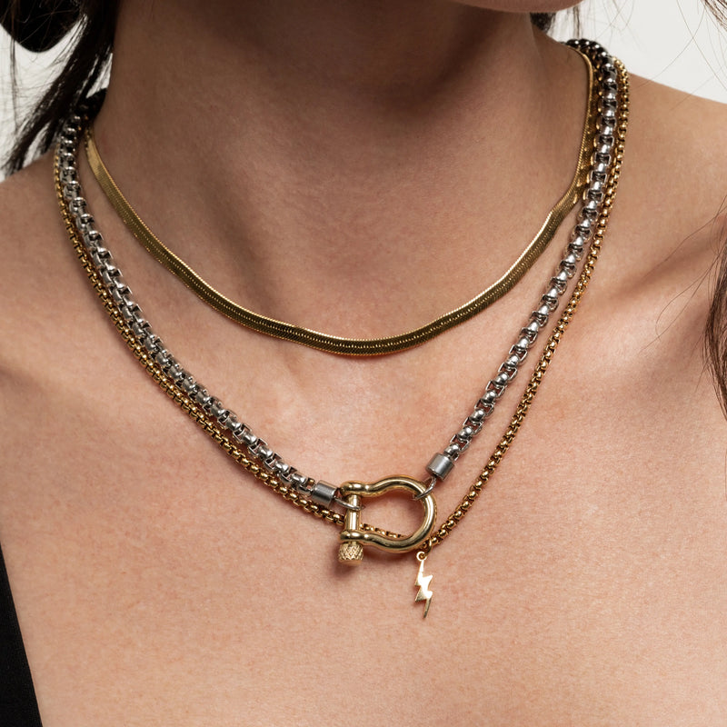 The Camaleon Layered Necklace Set, which is a three-piece layered necklace set featuring a gold flat chain, a silver and gold chain with a toggle clasp, and a gold chain with a lightning bolt charm, worn by a model in a black top.