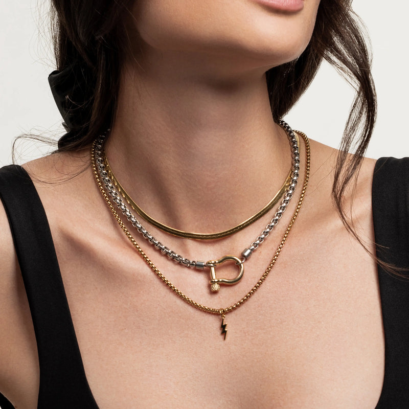 The Camaleon Layered Necklace Set, which is a layered necklace set featuring three necklaces: a gold and silver chain with a toggle clasp, a gold chain with a lightning bolt pendant, and a silver chain, worn by a woman in a black top.