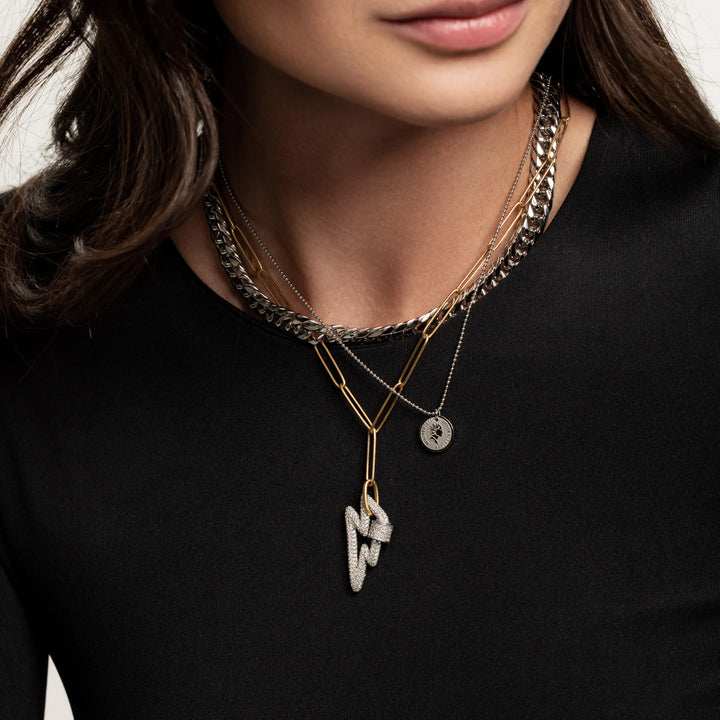 The Lightning layered necklace set, which is a three-piece layered necklace featuring a combination of silver and gold tones. It includes a chunky silver chain, a gold chain with a minimalist disc pendant, and a third chain with a striking lightning bolt pendant, creating a bold and dynamic look.
