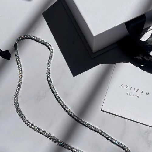 The image shows a silver necklace with diamonds. The necklace has a delicate chain with two rows of diamonds. The diamonds are small and appear to be white. The necklace is resting on a white surface next to a white gift box with a black ribbon. The gift box has the word 