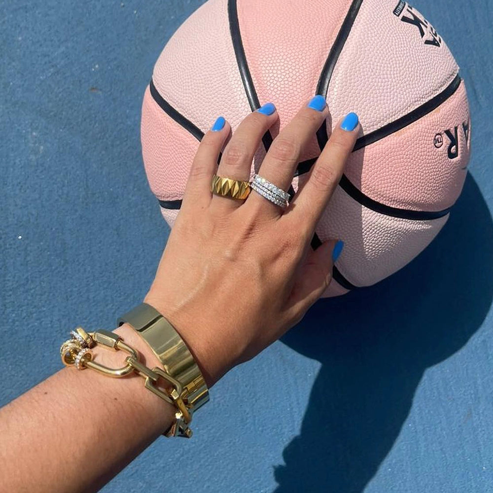 In the image, a woman is wearing a gold ring with diamonds, a gold chain bracelet, and has blue nail polish on. The ring appears to be a cocktail ring with a large, oval-shaped diamond in the center. The diamond is surrounded by smaller diamonds, which give the ring a sparkling effect. The chain bracelet is a thick, chunky bracelet with a gold clasp. The nail polish is a bright blue color.