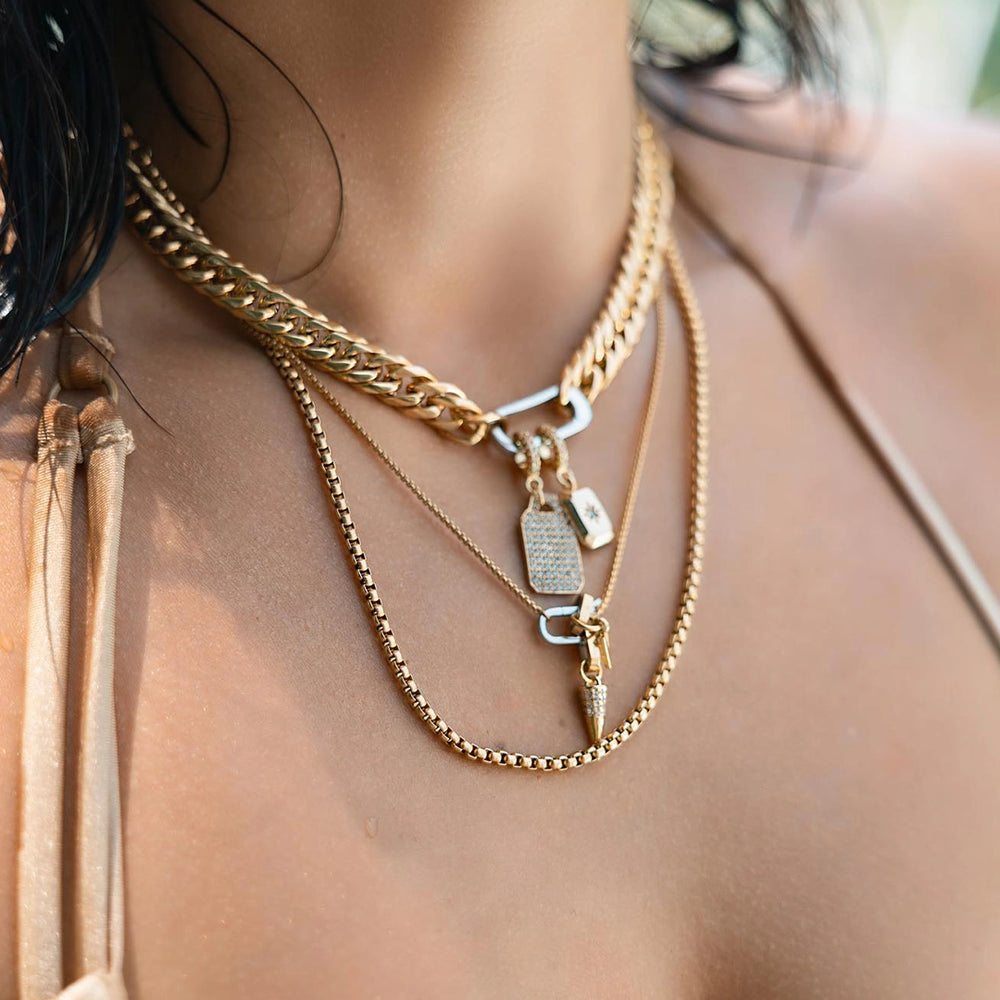 A woman wearing layered gold necklaces with an Artizan's JUST CLICK TAG NECKLACE SET chunky chain, a dog tag pendant, a diamond-encrusted heart pendant, and a spike-shaped pendant.