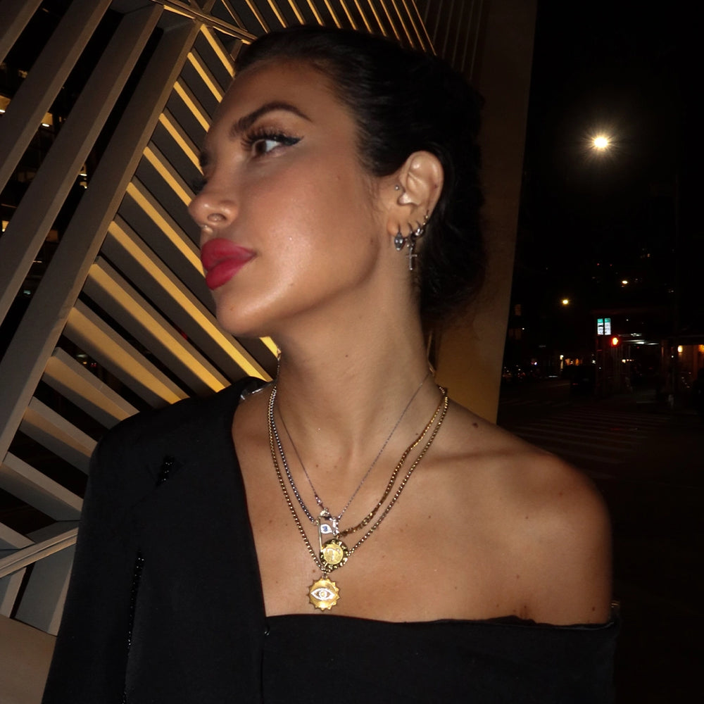 Side profile of a woman wearing layered gold and silver necklaces with eye and coin pendants, complemented by multiple earrings in one ear. She stands in an urban setting at night, with dramatic lighting highlighting her bold red lipstick and stylish black outfit.