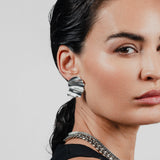 The WAVES OF SILVER EARRINGS, One piece, silver metal, bold shell-shaped earrings worn by model. Simple, elegant design with textured finish.