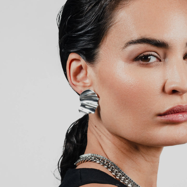 The WAVES OF SILVER EARRINGS, One piece, silver metal, bold shell-shaped earrings worn by model. Simple, elegant design with textured finish.