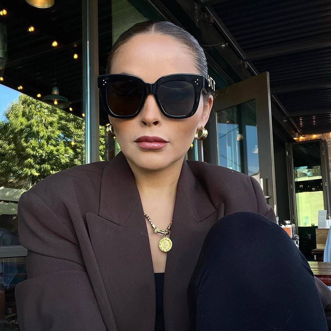 Stylish woman wearing oversized black sunglasses, gold hoop earrings, a chunky gold necklace with a medallion pendant, and a brown blazer, posing outside a modern café.
