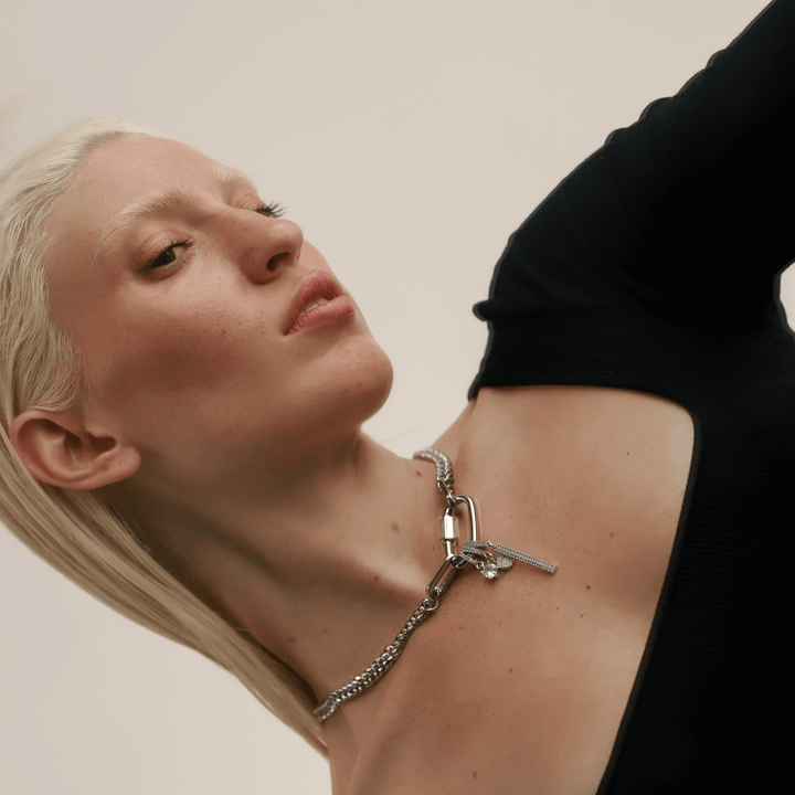 Model wearing the ANNE CLIP ON SHINE NECKLACE: A bold and modern necklace showcasing a thick mixed-metal chain in silver and gold tones. The focal point is a gold carabiner clasp adorned with a diamond-encrusted circular pendant and a miniature padlock charm, adding a touch of sparkle and playful sophistication.
