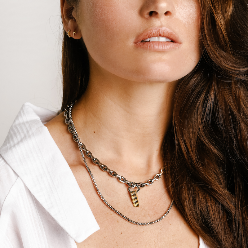 Model wearing the SILENCE LAYERED NECKLACE SET which comes in 3 separate chains. It includes a stainless steel puerto chain which is 17 inches in length with gold triangle charms & Zirconia, one thin gold plated necklace and another thin silver necklace that are 16 and 20 inches in length. She is also wearing a mini lightning earring and the huggies earring.