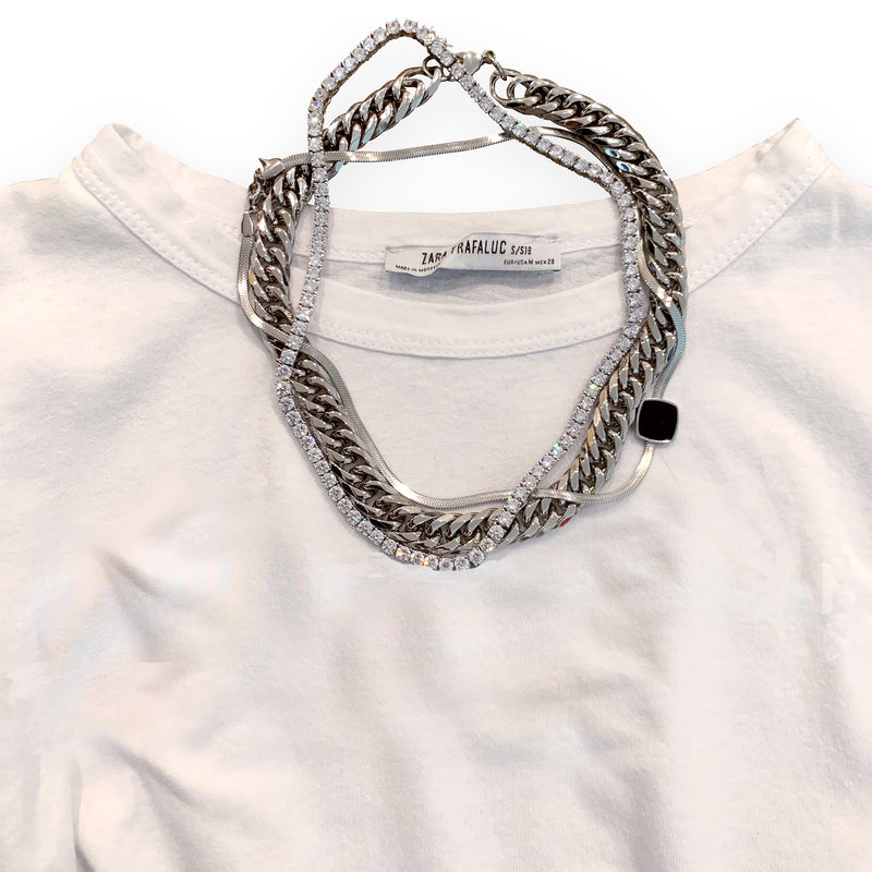 Synergy Set which comes with two necklaces. One is the palma necklace which is a silver chain with box type lock and another one is the tennis necklace made of Clear Zirconia Stones. It is layered with the Square snake in silver necklace placed on top of a white tshirt.