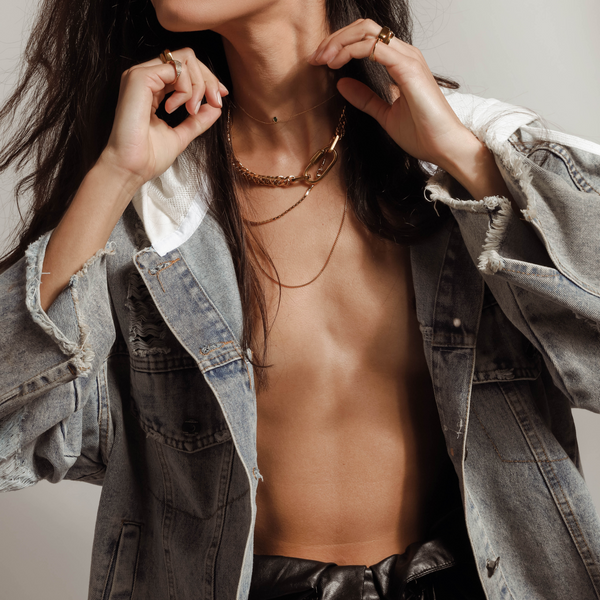 Model wearing an oversized denim jacket and The ANNE GOLD NECKLACE SET which includes 4 separate layered chains. The shortest is the Anne Necklace made of chunky 18k gold plated stainless steel chain with carabiner clasp followed with two thin necklaces that are 20" and 24" in length and a necklace with 925 Sterling Silver Gold plated emerald drop dainty charm.