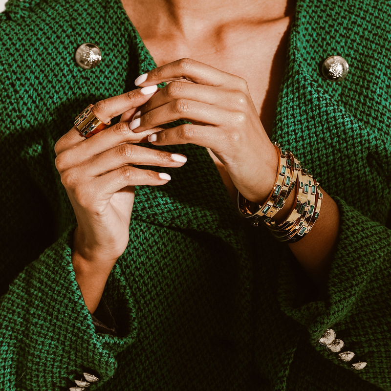 Model wearing a stack of Emerald Bangle and Emerald thin bangles. She is wearing three emerald rings that are gold plated stainless steel with three emerald stones encrusted. .