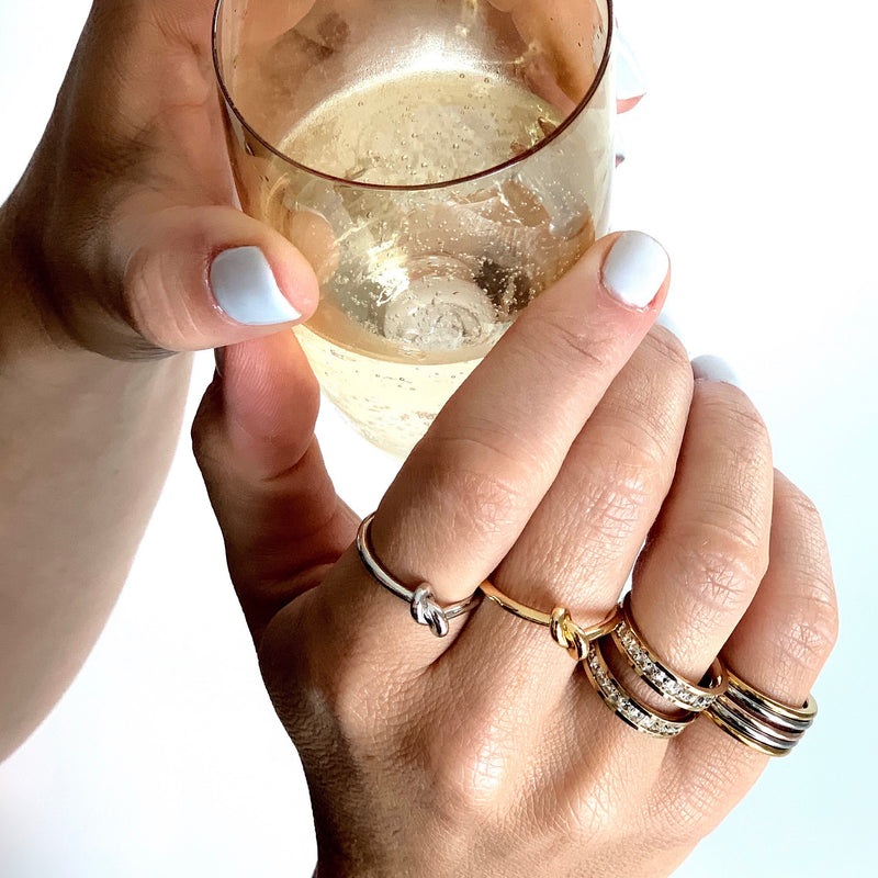 Model's hand holding a Champagne glass wearing the knot rings in silver and gold, square rings in silver and gold.