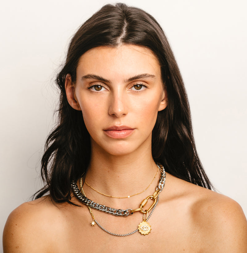 Model wearing the Amelia Layered Necklace Set Mix. Shortest is a thin gold necklace next to a chunky silver chain with gold oval clasps and Gold Eye Pendant. The longest is a half gold half silver thin chain with a small gold star pendant.